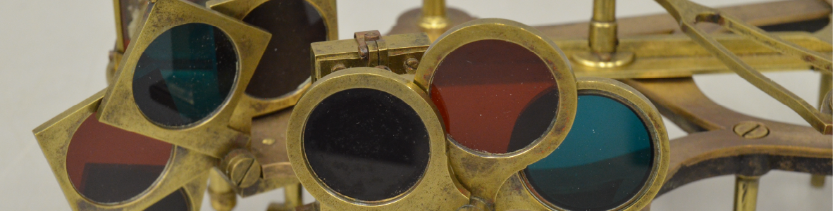 Close-up of an artifact, indistinguishable aside from two sets of three yellow-bronze frames holding circular, translucent lenses. The lenses are red, blue, and black, with one set in square frames and the other in circular frames. The rest of the artifact's bronze apparatus can be seen out of focus in the background.